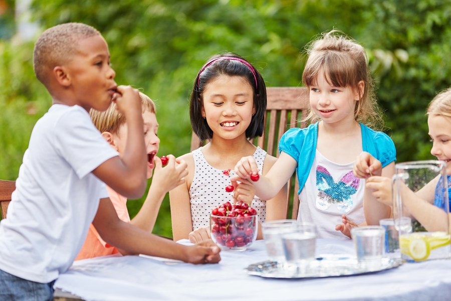 Das Foto zeigt Kinder im Garten. Sie sitzen lachend am Tisch und essen Kirschen. 