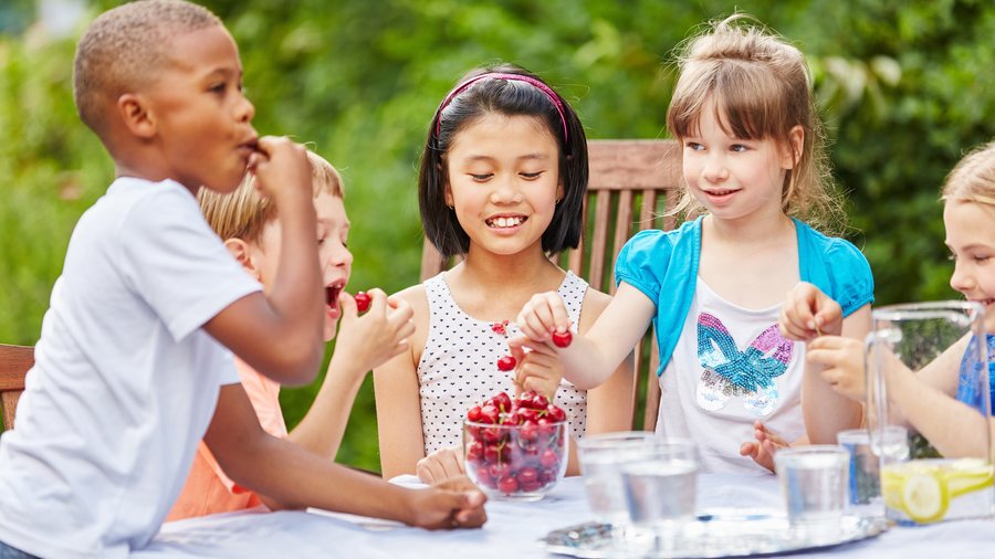 Das Foto zeigt Kinder im Garten. Sie sitzen lachend am Tisch und essen Kirschen. 